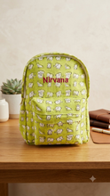 Load image into Gallery viewer, A light green backpack with a repeating white cartoon elephant pattern and the word 'Nirvana' in red across the front, sitting on a wooden table with a small plant and notebook nearby.